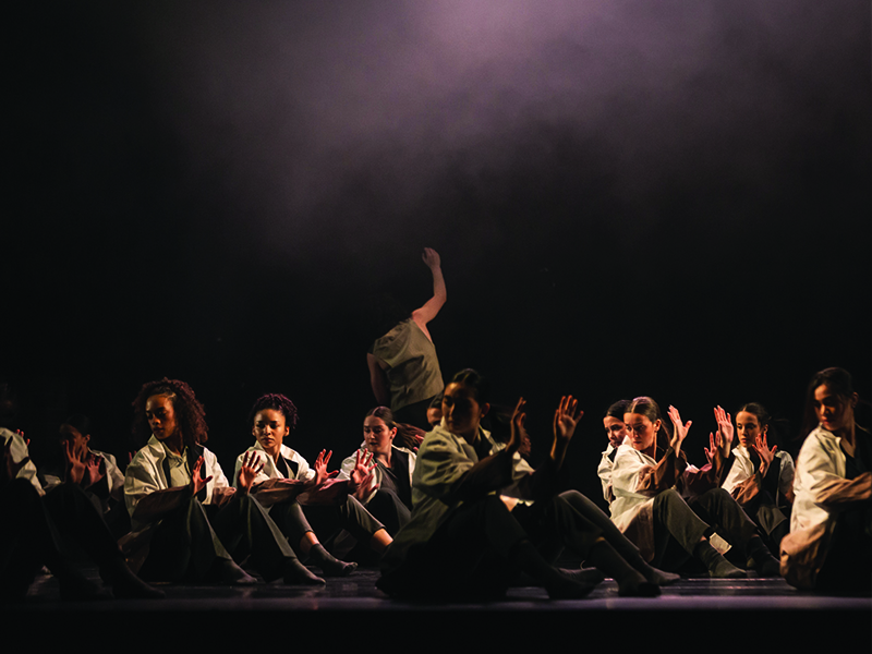 Dancers are seated on stage while performing while one dancer is standing at the back of the stage